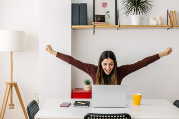 Excited young woman celebrating good news working on laptop computer in the living room at home....