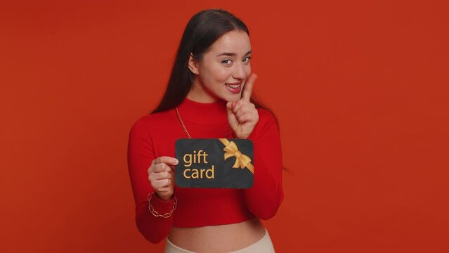 Young woman in crop top showing pointing on gift discount certificate voucher coupon for store holidays sale. Lovely girl with shopping christmas surprise card isolated alone on red studio background