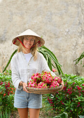 Positive adult female in white shirt and conical hat standing in garden and carrying wicker basket with dragon fruits