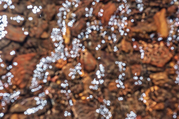 Abstract blur nature background of clear water stream with motley stony bottom in sunlight. Defocused mountain stream texture in bright sun. Blurry pattern of sunlit multicolor stones in spring water.