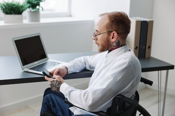 A man wheelchair in the office works at a laptop with a phone, working online, social networks and startup, integration into society, the concept working person with disabilities, real person close-up