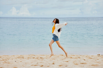 A woman runs along the beach in summer clothes on the sand in a yellow T-shirt and denim shorts white shirt flying hair ocean view
