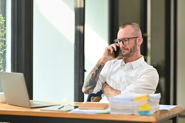 Confident handsome businessman in a white shirt with tattooed talking mobile phone and using laptop at his workplace