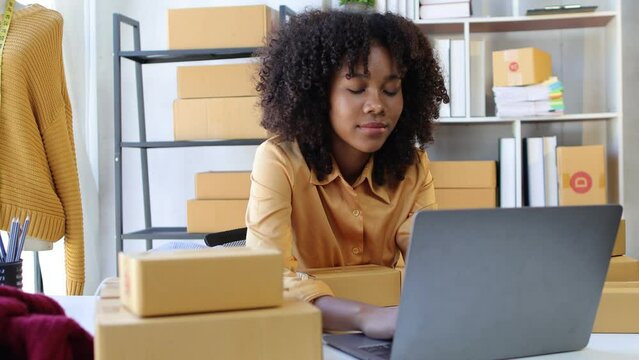 African young woman owner of a start up business designing and selling clothes online checking orders on laptop and preparing for delivery.