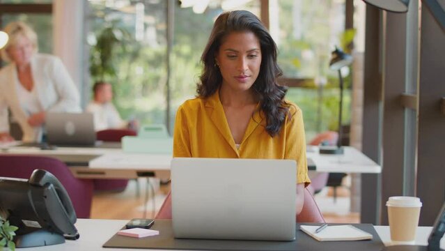 Mature businesswoman working on laptop at desk in modern office pauses to look out of the window and smile - shot in slow motion