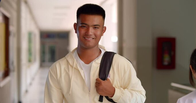 Slow motion scene of a happy smiling male high school student standing on a corridor in a school building with a laptop backpack.