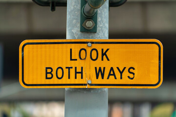 Orange yellow road sign that say look both ways with black letters on side of street light with metal and blurry uraban