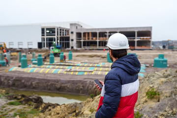 Engineer builder in a hard hat with a smartphone on the background of the construction of an industrial building