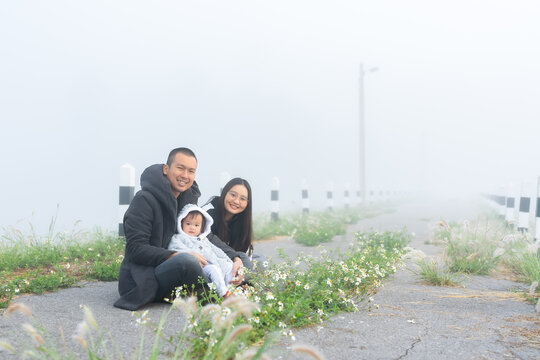 happy family in morning with cold weather and fog covering area, father mother take their cute little daughter come see fog feel thick air, mother giving flowers to play with daughter.