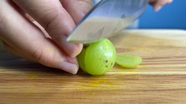 Female hands using knife to cut raw indian gooseberry slice on a wooden chopping board at home kitchen close up shot. Healthy lifestyle, fresh vegetable and cooking concept