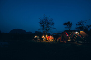 Asian tourists camping at night on the coast of Laos tourist destination in front of a tent at a campfire under the starry evening sky and the Milky Way on blue water and forest floor. Outdoor tent li