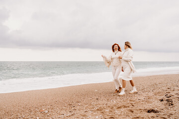 Fototapeta na wymiar Women sea walk friendship spring. Two girlfriends, redhead and blonde, middle-aged walk along the sandy beach of the sea, dressed in white clothes. Against the backdrop of a cloudy sky and the winter 