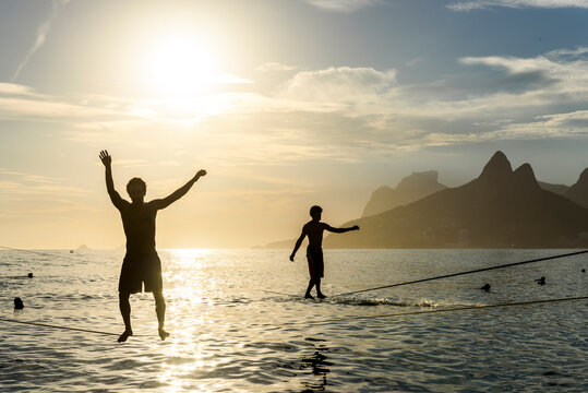 Silhouette Of Two Men Balances On A Slackline At Arpoador Beach During The Sunset, Rio De Janeiro, Brazil