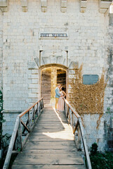 Groom hugs bride leaning against the railing of the wooden bridge to Mamula Fort