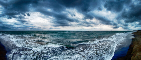 Dramatic weather panoramic view on the beach with overcast sky and nimbostratus clouds and stormy sea waves crashing at the shore with many sea foam