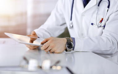 Unknown male doctor sitting and working with tablet computer iin a darkened clinic, glare of light on the background, close-up of hands