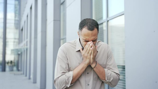 Young adult man sneezes because he has seasonal allergies while standing outside an office building. Male has the first symptoms of cold and flu, pneumonia, bronchitis and respiratory tract infection