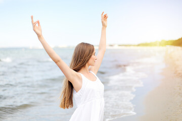 Fototapeta na wymiar Happy smiling woman in free happiness bliss on ocean beach standing with raising hands. Portrait of a multicultural female model in white summer dress enjoying nature