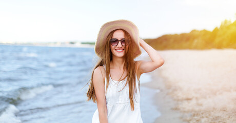 Fototapeta na wymiar Happy smiling woman in free happiness bliss on ocean beach standing and posing with hat and sunglasses. Portrait of a female model in white summer dress enjoying nature