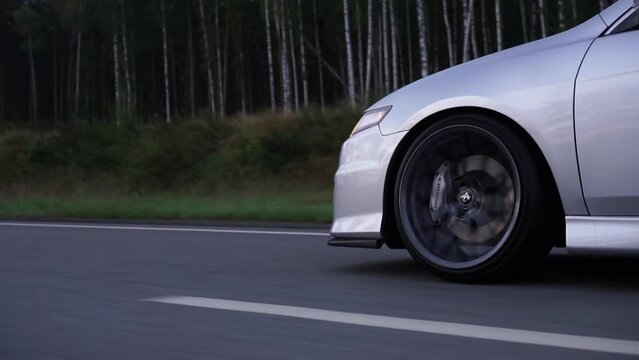 Side view of silver car rides on summer highway. Footage. Perfect auto drives fast on country road. Big sports alloy wheels with powerful brakes