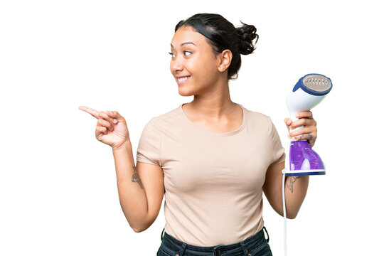 Young Uruguayan woman holding a vertical steam iron over isolated chroma key background pointing to the side to present a product