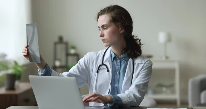 Woman radiologist, medical worker in uniform sits at workplace with laptop, holding digital image, analyze x-ray, reviewing patient health state result. Healthcare, radiology, professional occupation