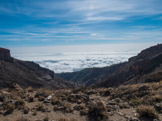 View from mountain pass of Alto de Guajara volcano with with lava rock boulders and dry vegetation, above the white clouds, blue sky background. El Teide National Park, Tenerife, Canary islands, Spain