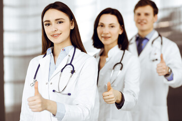 Fototapeta premium Group of young cheerful doctors is standing as a team with thumbs up in a hospital office and is ready to help patients. Medical help, insurance in health care