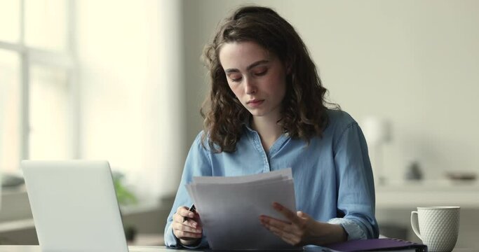 Young 20s thoughtful woman working with papers or studying, reading essay, learning, check written information, prepare assignment, making test, looks concentrated sits at desk with laptop. Paperwork