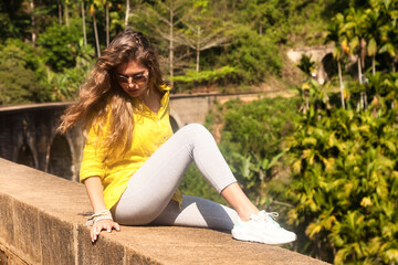 Young woman in sunglasses sitting on nine arch railway bridge at sight landmark background. Happy lady looking down enjoying at tropical journey; Ella; Sri Lanka. Travel vacation concept. Copy space