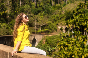 Happy woman in sunglasses sitting on nine arch railway bridge at sight landmark background. Happy lady looking away enjoying at tropical journey, Ella, Sri Lanka. Travel vacation concept. Copy space