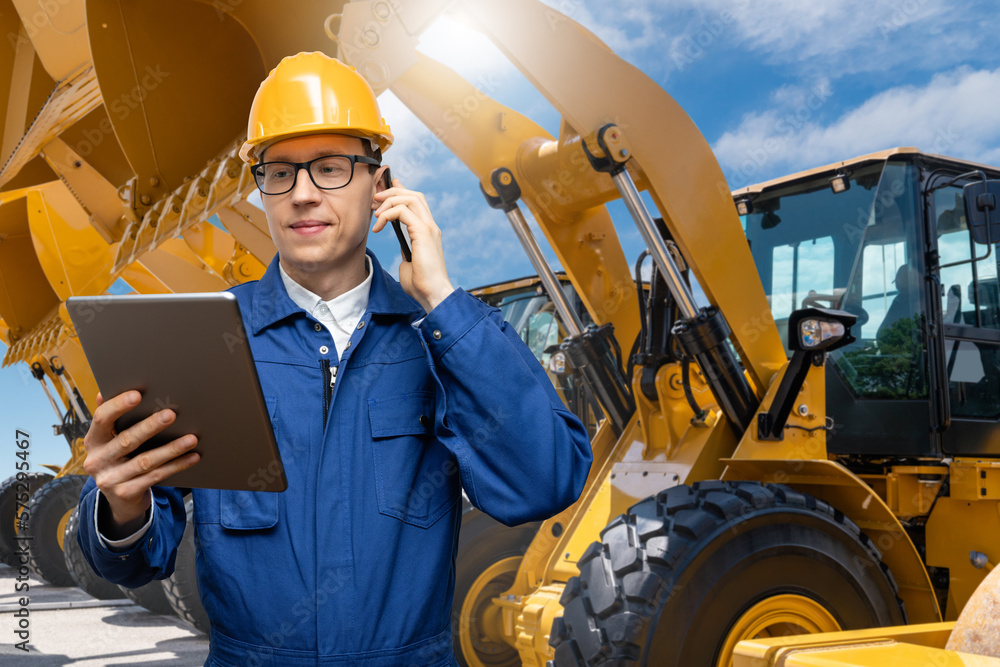 Aufkleber Engineer in a helmet with a digital tablet and phone on the background of construction machines	