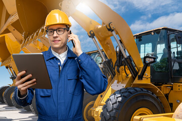 Engineer in a helmet with a digital tablet and phone on the background of construction machines	