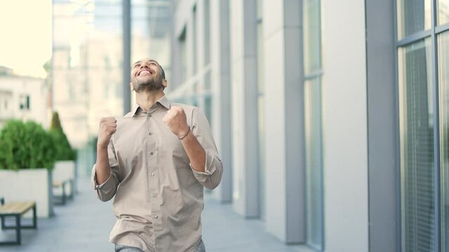 An excited adult young man celebrates victory, walking the street near the office building. A happy beautiful businessman jumps, dances, showing her hands a triumphant gesture. The concept of success
