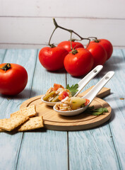  Appetizers of potato salad, tomato, green beans, egg and tuna in ceramic spoons on a wooden board with a bunch of ripe tomatoes.