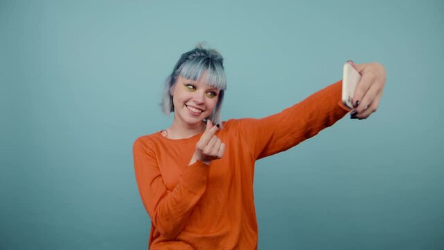 Positive teenage girl with colored hair taking selfie on blue background and showing heart gesture.