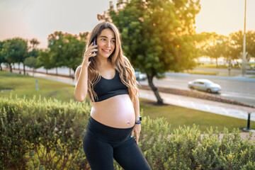 Portrait of smiling pregnant woman in sportswear talking on phone in a park. Pregnancy, technology and healthy lifestyle concept.