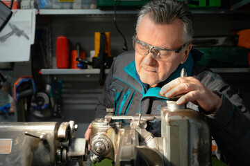 A man in safety glasses and a work uniform sets up a grinding machine in his workshop. The grinding machine details in focus. In the background are various working tools on the metal shelving for