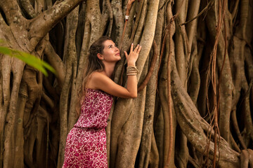 Young cute lady posing on background of wood branches or lianas in tropical. Pretty traveler woman cuddling trunk of large tree, looking up. Travel vacation and ecology concept. Copy ad text space