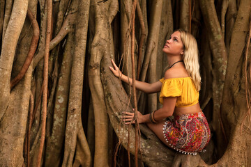 Young cute lady sitting on background of wood branches or lianas in rainforest, looking up. Pretty traveler woman cuddling trunk of large tree. Travel vacation and ecological concept. Copy text space - obrazy, fototapety, plakaty