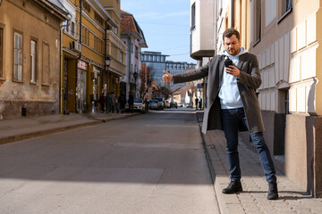 Man in coat standing on curbside hailing a taxi and looking at his cell phone.