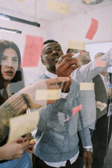 A front view photo though glass wall of two multiracial colleagues writing on sticky notes over...