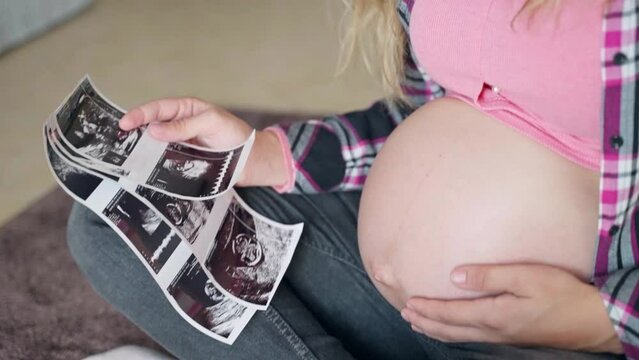Necessary medical examination of pregnant woman before childbirth, pregnant woman sitting on the floor, holding ultrasound scan pictures and stroking gently her beautiful pregnant belly. Preparation