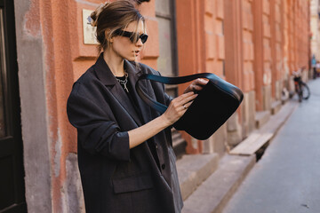 Elegant young woman looking in her black leather bag her phone or purse. Business style woman wear...