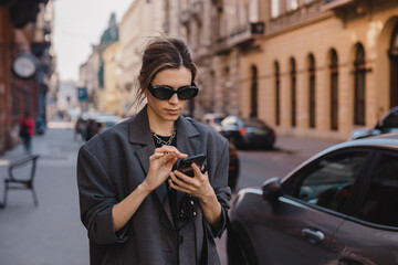 Fototapeta premium Young stylish business woman writes message on smartphone walking on old city street, hipster tourist traveling in european town and planning trip using mobile phone.