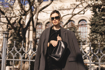 Fototapeta na wymiar Elegant young woman looking in her black leather bag her phone or purse. Business style woman wear grey blazer, black eyeglasses and bag on the street. Street style, fashion outfit.
