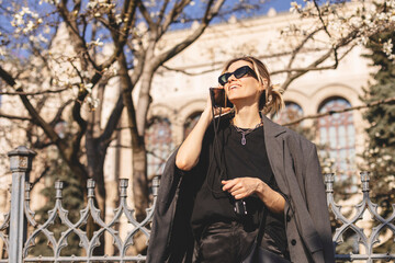 Beautiful young woman wearing suit, walking outdoors, talking on mobile phone. Beautiful Woman talking on phone. Portrait of stylish smiling business woman calling on mobile phone under blooming tree.