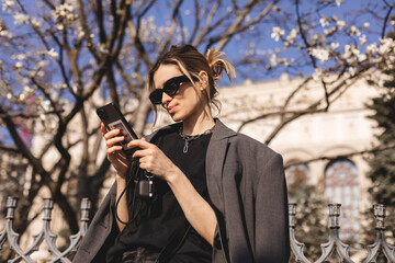 Happy woman walking using smart phone in a city street. Business woman with sunglasses uses mobile phone outdoors under blooming tree, girl typing message, chatting, scrolling web page, read good news