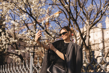 Fototapeta na wymiar Happy woman walking using smart phone in a city street. Business woman with sunglasses uses mobile phone outdoors under blooming tree, girl typing message, chatting, scrolling web page, look at camera