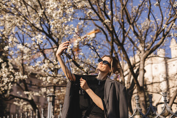 Fototapeta na wymiar Happy woman walking using smart phone in a city street. Business woman with sunglasses uses mobile phone outdoors under blooming tree, girl typing message, chatting, scrolling web page, look at camera
